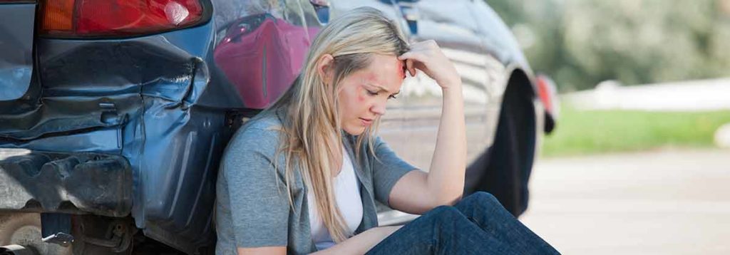 A bruised woman sitting next to her damaged vehicle after a car accident. She is waiting to speak with a personal injury lawyer.