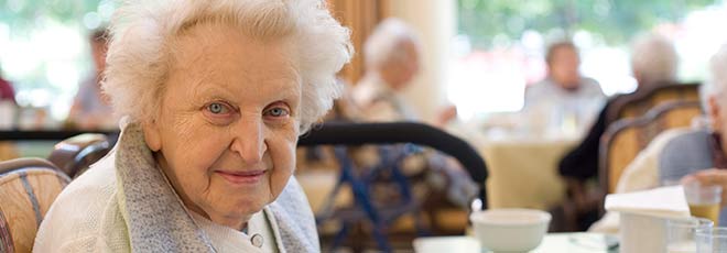 An older woman smiles at the camera while sitting at a table in a nursing home.