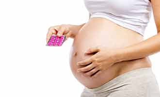 The focus is on the belly of a pregnant individual as she holds pills in her hand.