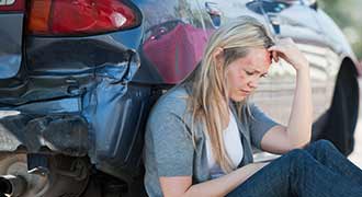 A bruised woman sitting next to her damaged vehicle after a car accident. 