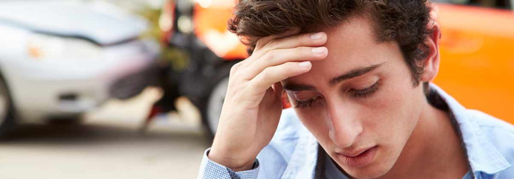 A teenager holds his head in distress after being in a car accident. The two vehicles are damaged in the background.