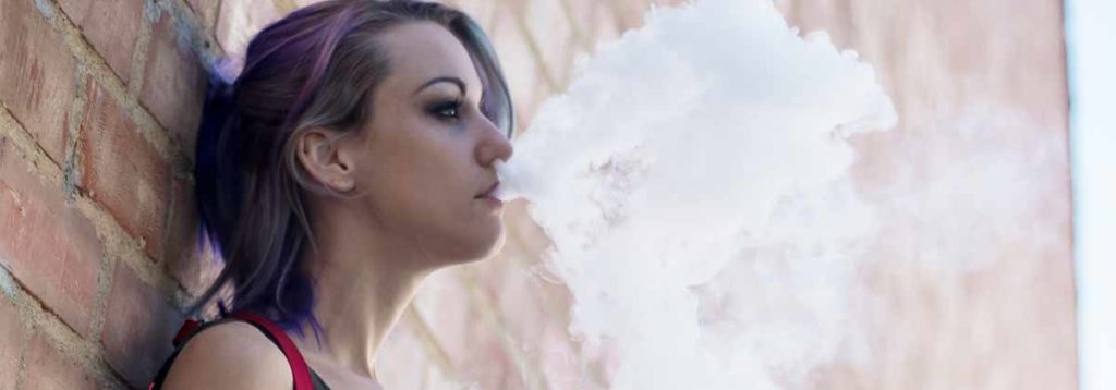 A woman with her back to thew wall while vaping. 