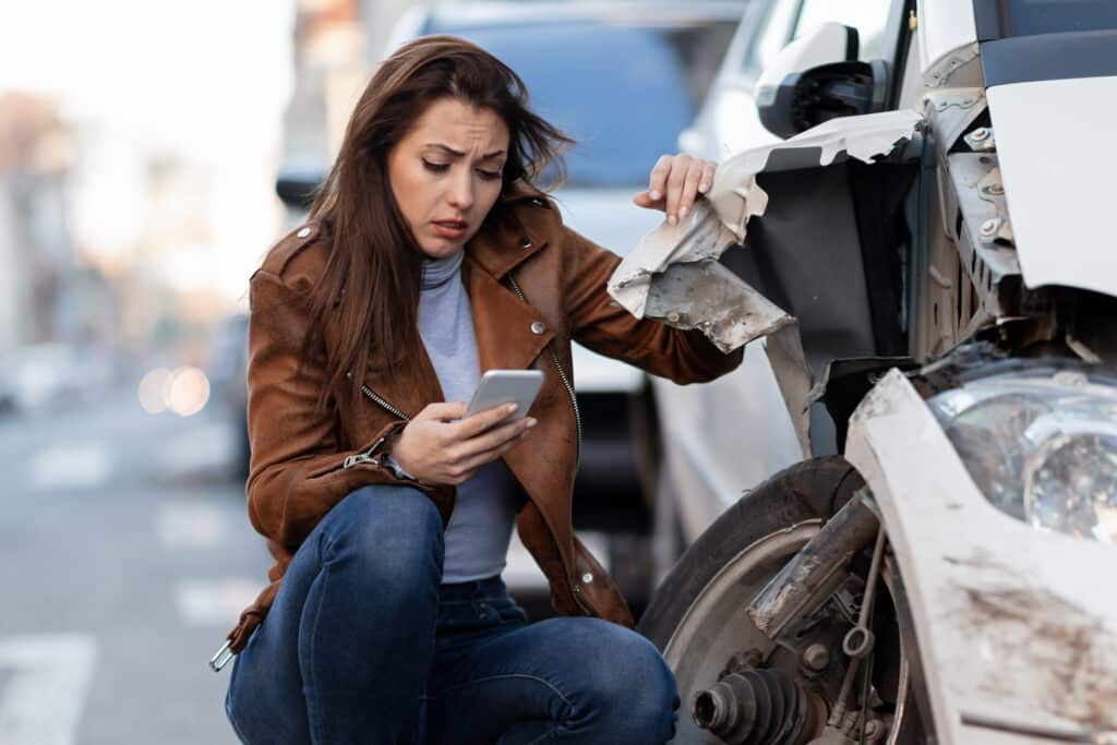 Young woman texting a Massachusetts auto accident lawyer while inspecting her damaged car
